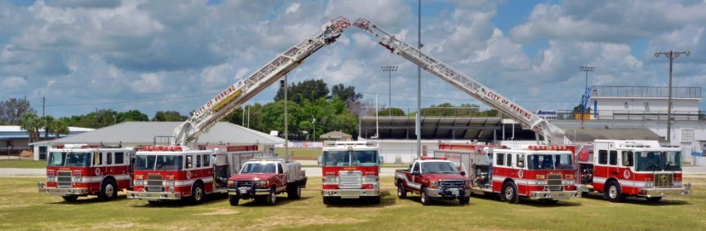 7 fire trucks lined up at fireman's field with both ladder trucks extended towards each other